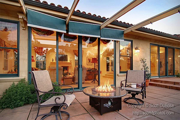 Outdoor patio with fire pit table, wrought-iron lounge chairs, retractable awning, and glass sliding doors at dusk photographed by Scott DuBose