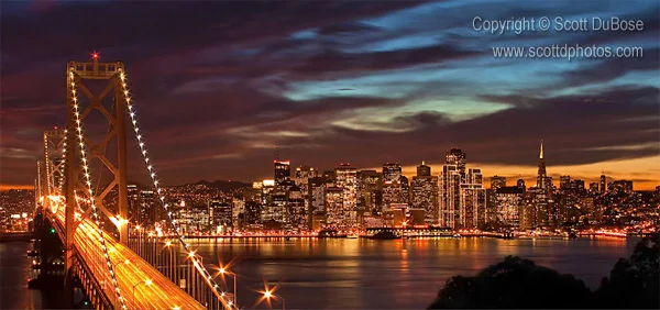 Night panorama of the San Francisco Bay Bridge with the illuminated city skyline reflected in the water photographed by Scott DuBose