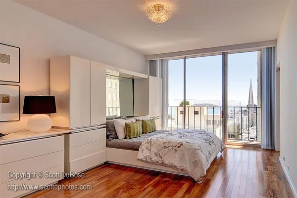 Modern urban bedroom with floor-to-ceiling windows, city skyline view, white built-in wardrobes, and hardwood floors photographed by Scott DuBose