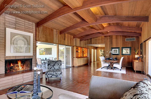 Mid-century open-plan living and dining room with wood beam ceiling, lit fireplace, and terracotta tile floor photographed by Scott DuBose