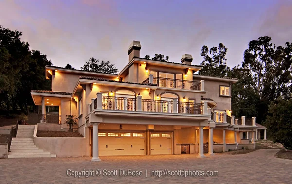 Night exterior of a luxury multi-story home with illuminated balconies, triple garage, and stone driveway photographed by Scott DuBose